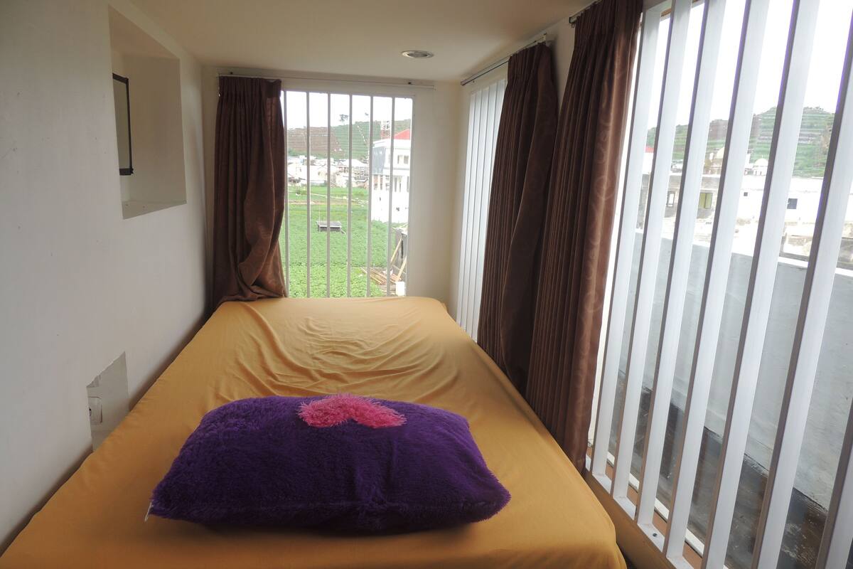 Uma cama de solteiro com colcha amarela e travesseiro roxo, uma janela com cortinas, e vista para uma paisagem verde lá fora.