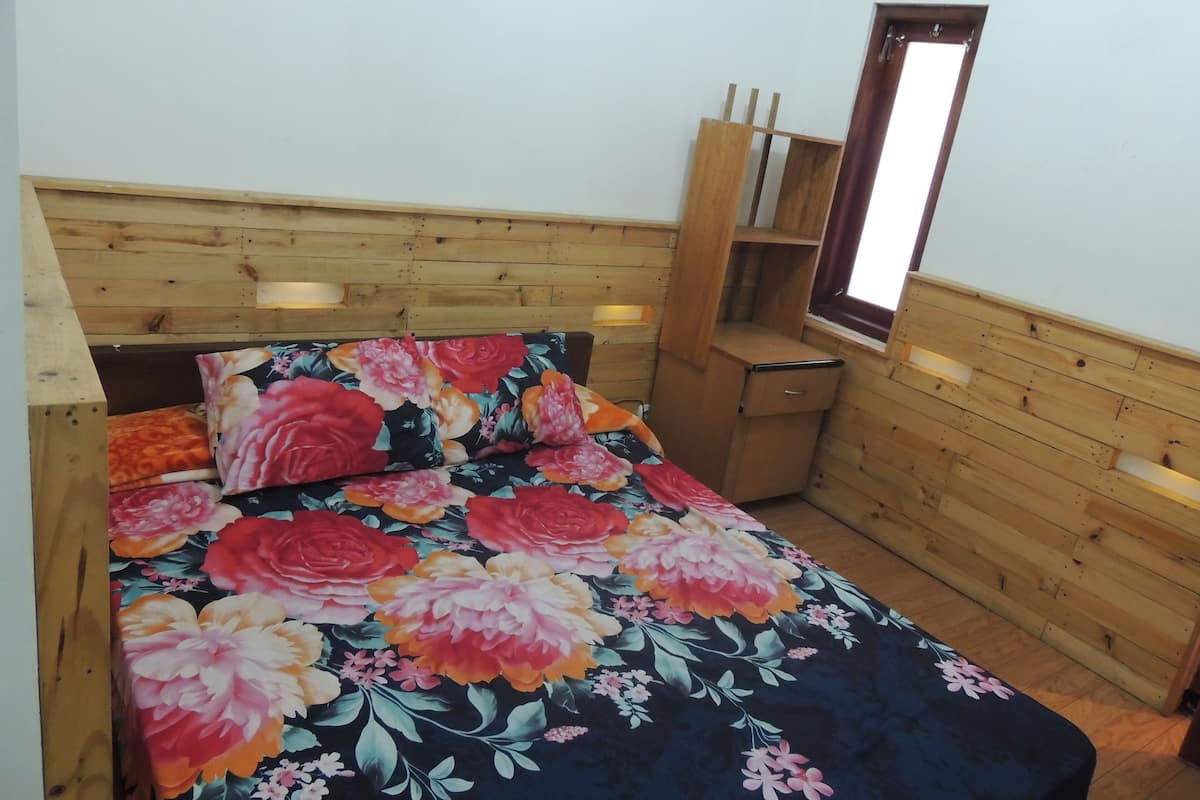 A small, cozy room with a wooden headboard, a bed with a floral bedspread, a nightstand, a wooden dresser, and a window with a view.