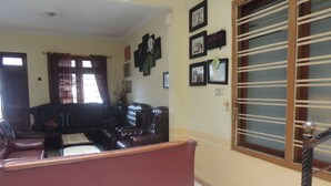 Una sala de estar con un sofá marrón oscuro, un sofá rojo y una mesa de centro. Hay cuadros en la pared y una ventana con persianas.