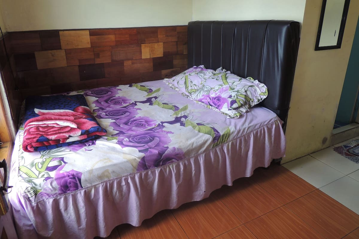 A bed with a floral bedspread, a headboard with a wooden panel design, and a plain wall with a mirror.