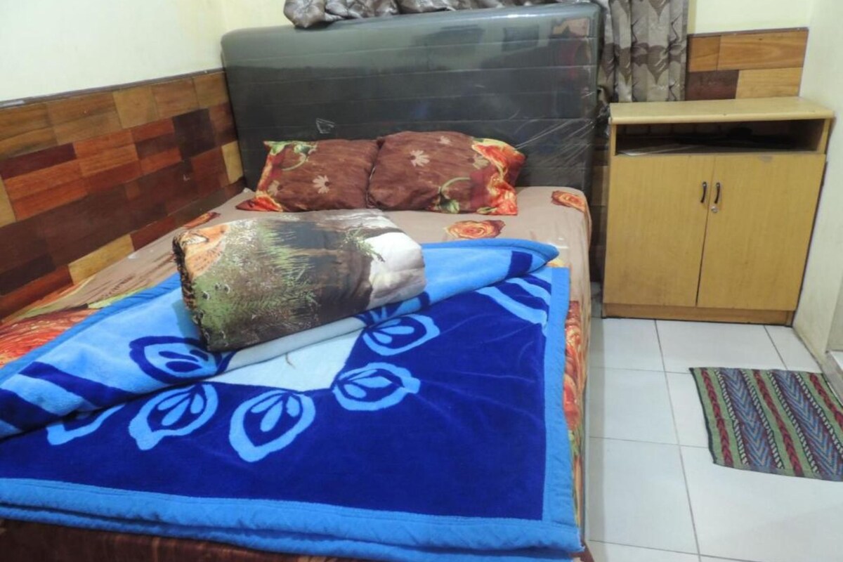 A bed with a blue blanket and a decorative pillow, a wooden headboard, a small wooden cabinet, and a patterned rug on a tiled floor.