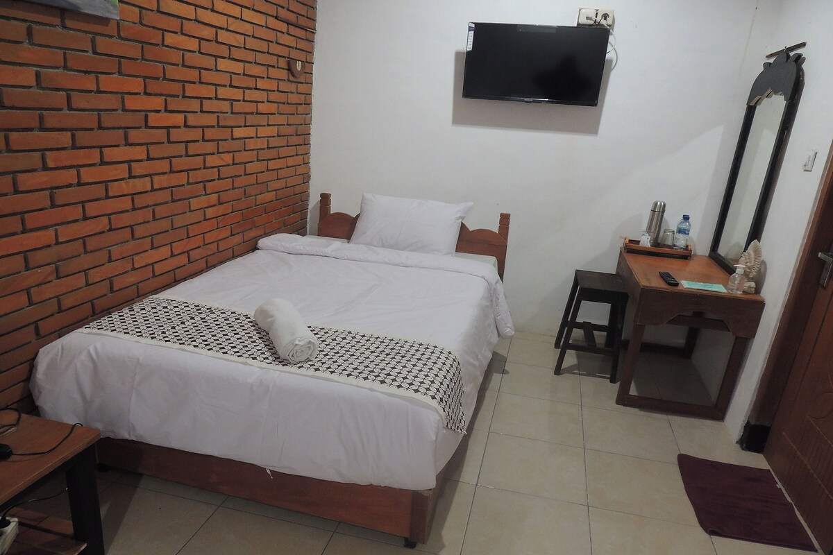 Una habitación de hotel con pared de ladrillo, cama con ropa blanca, escritorio con espejo y televisor montado en la pared.