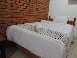 Una cama con ropa blanca y una manta estampada, cabecera de madera y pared de ladrillo.