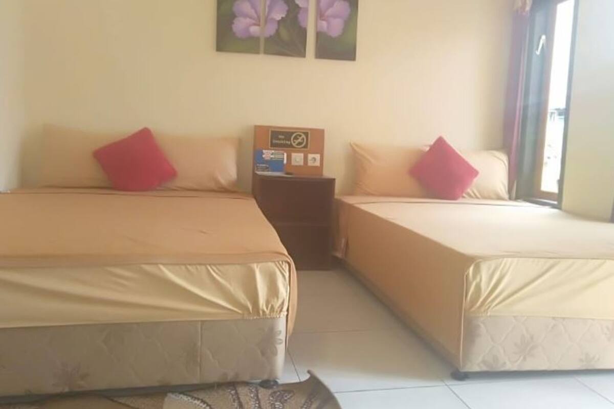 Quarto de hotel com duas camas, cada uma com colcha bege e travesseiro vermelho. Há uma mesinha com uma caixa em cima, e um quadro de flores na parede.