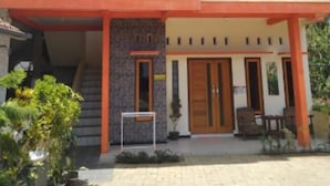 Il s’agit d’un immeuble à deux étages, sur lequel on peut lire l’inscription « KIKI HOMESTAY ». On y trouve plusieurs portes, un escalier et un petit espace extérieur aménagé avec des tables et des chaises.