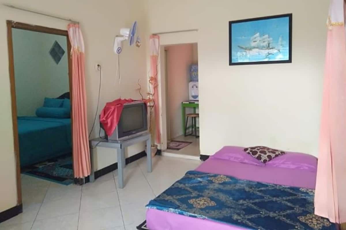 Um quarto com uma cama, uma televisão sobre um suporte, uma escrivaninha com uma cadeira e um quadro na parede.
