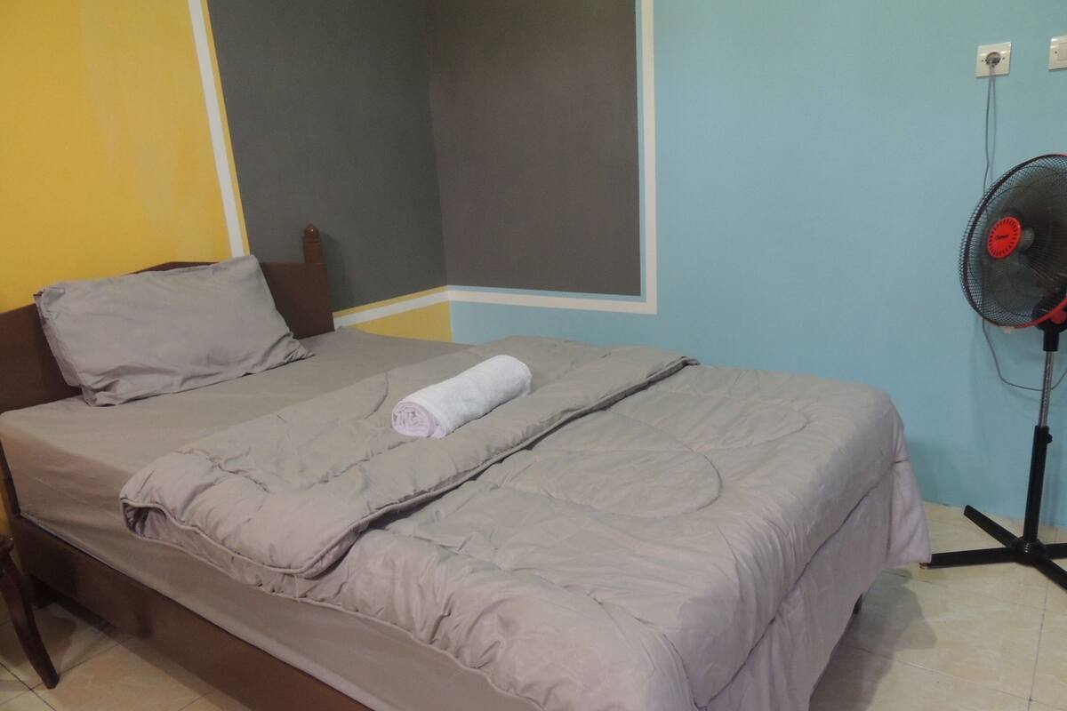 A single bed with a grey duvet, a rolled towel, and a grey pillow in a room with light blue walls and a grey floor.