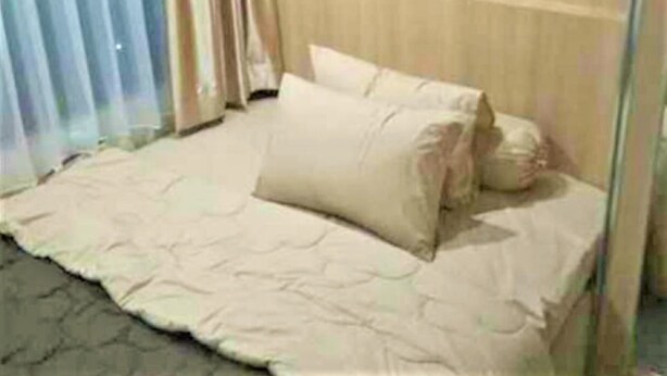 Un lit avec une couvre-lit et des oreillers de couleur beige, une tête de lit en bois et une fenêtre avec des rideaux.