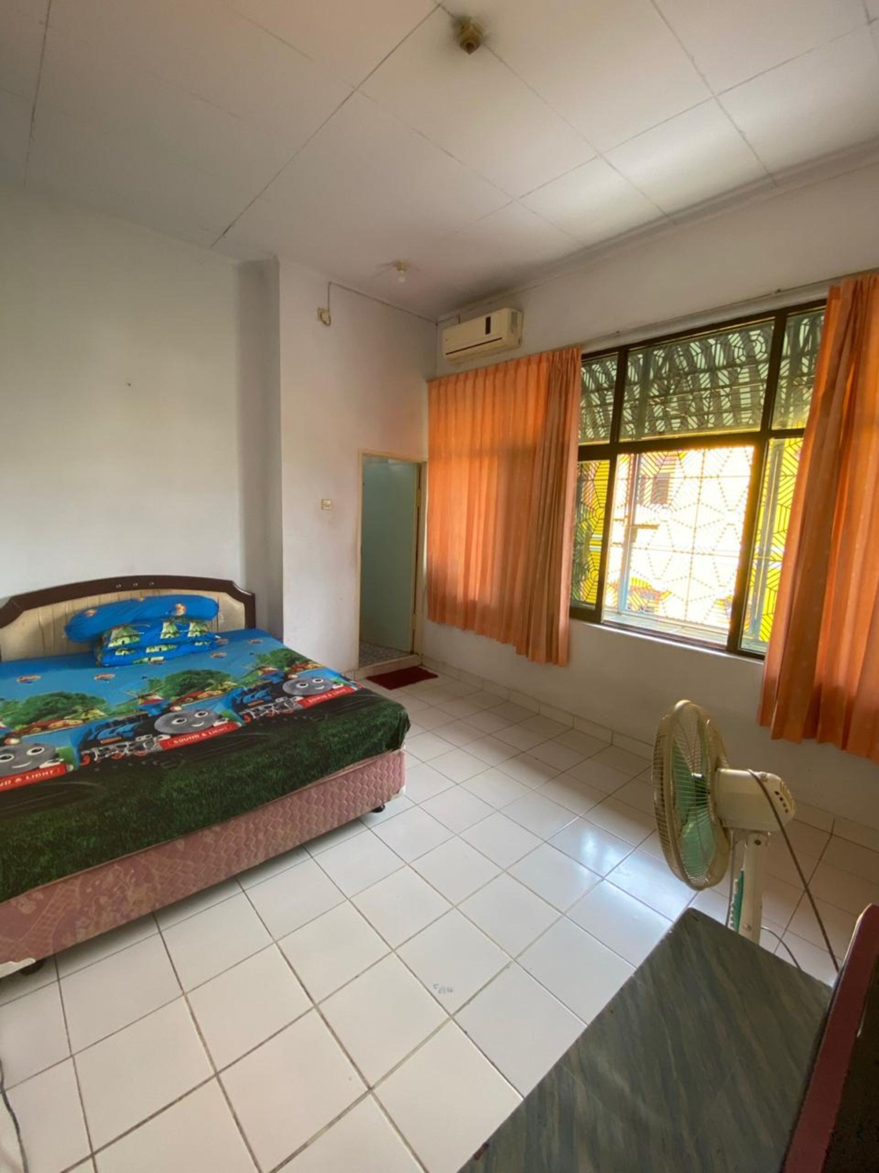 A bedroom with a bed, a window with curtains, a standing fan, and a door.