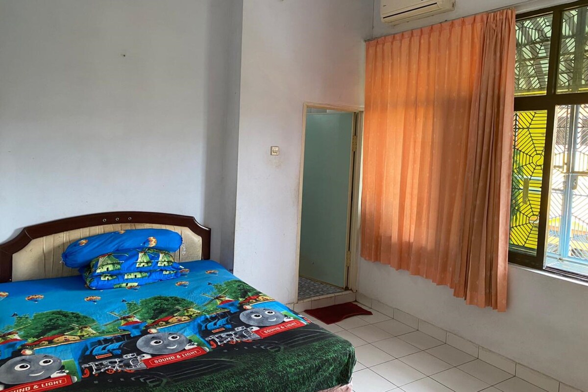 A bedroom with a bed covered in a Thomas the Tank Engine themed bedspread, a window with orange curtains, and an air conditioning unit on the wall.