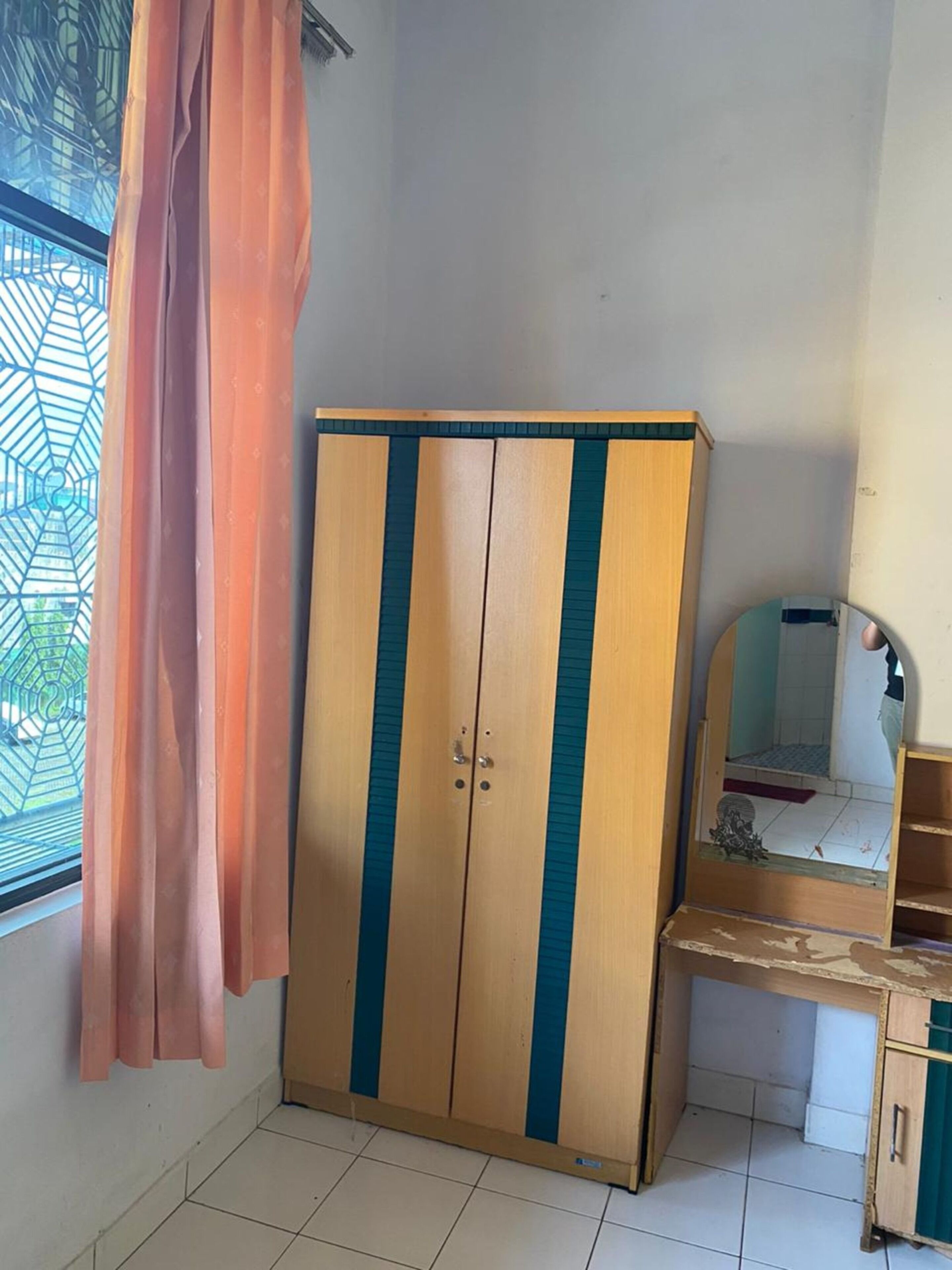 A room with a wooden wardrobe with blue stripes, a wooden desk with a mirror, and a window with a geometric pattern.