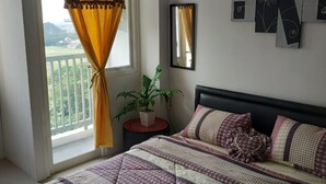 A bedroom with a bed, a potted plant, a nightstand, and a window with curtains. - Grand Sentraland Karawang by Gkit Room (Karawang)