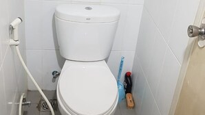 A small, clean bathroom with white tiles, a toilet, a handheld showerhead, and a blue cleaning brush. - Grand Sentraland Karawang by Gkit Room (Karawang)