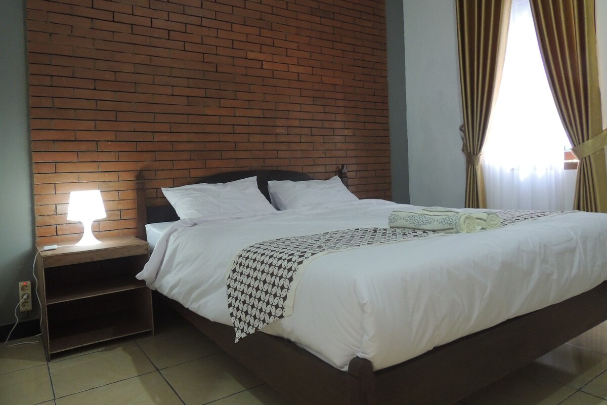 A bedroom with a brick wall, a bed with white bedding, a nightstand with a lamp, and a window with curtains.