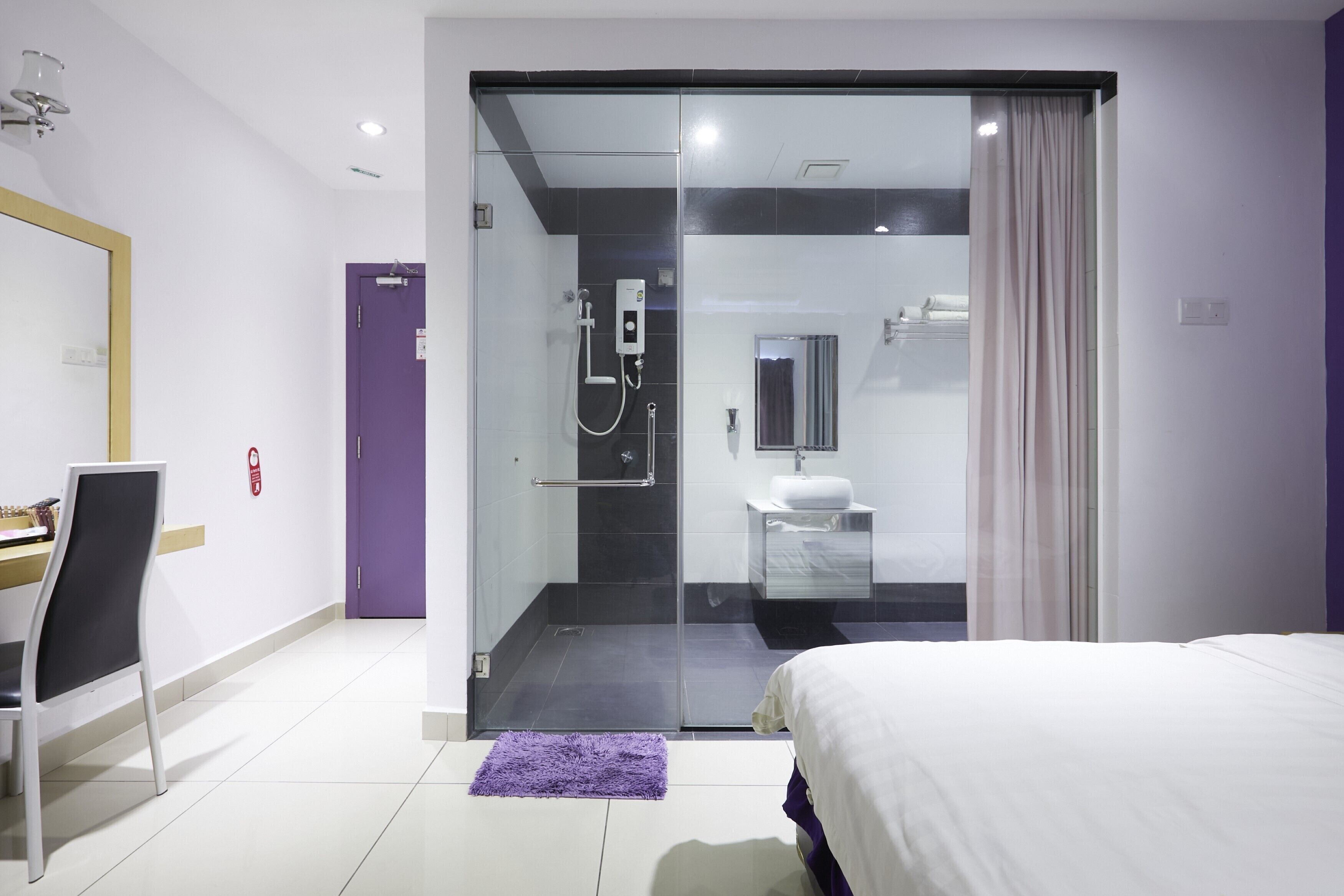 Et moderne hotelværelse med en brusekabine i glas, en seng, et skrivebord med stol og en lilla måtte.