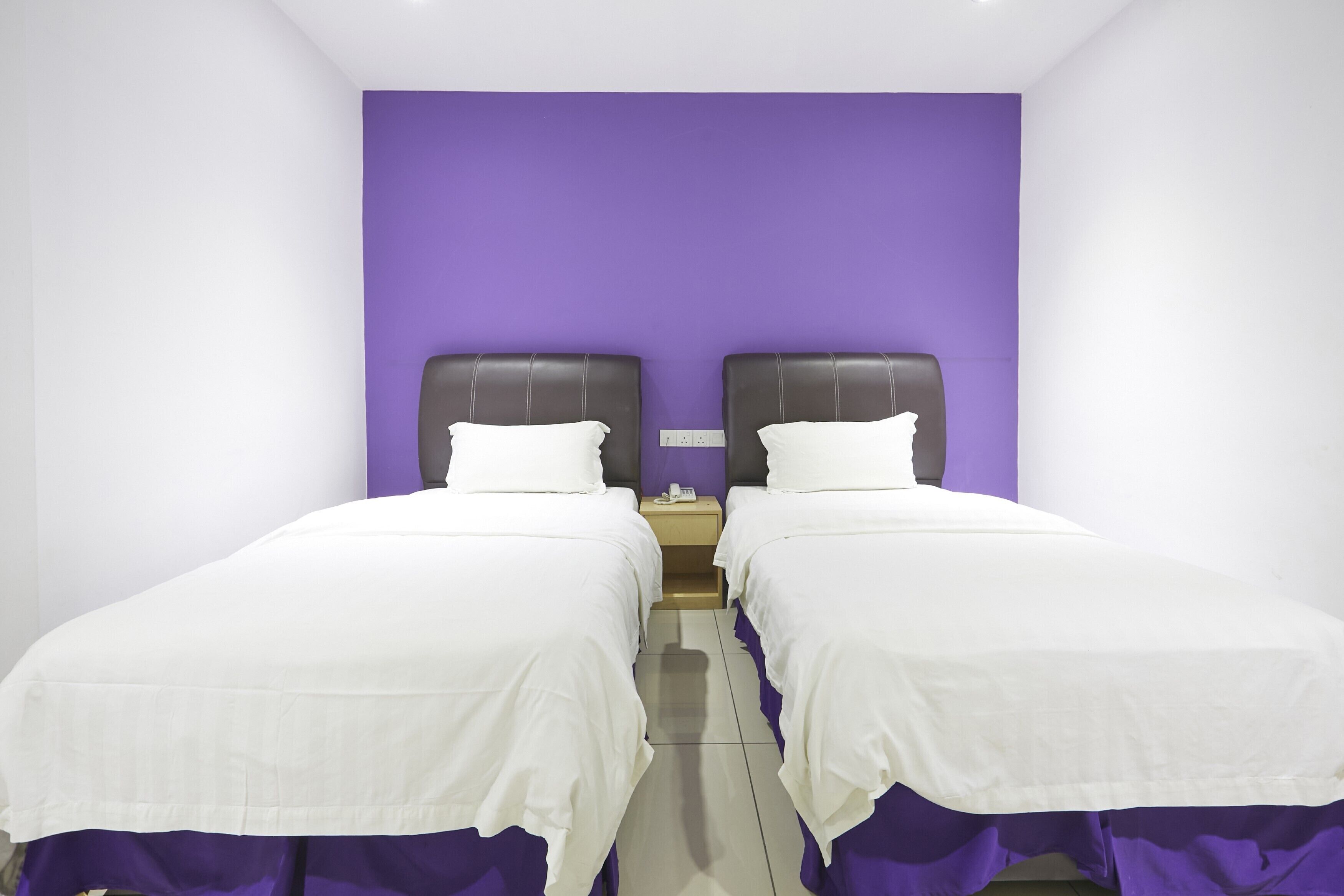 A room with two beds, each with a white bedspread and a purple wall behind.