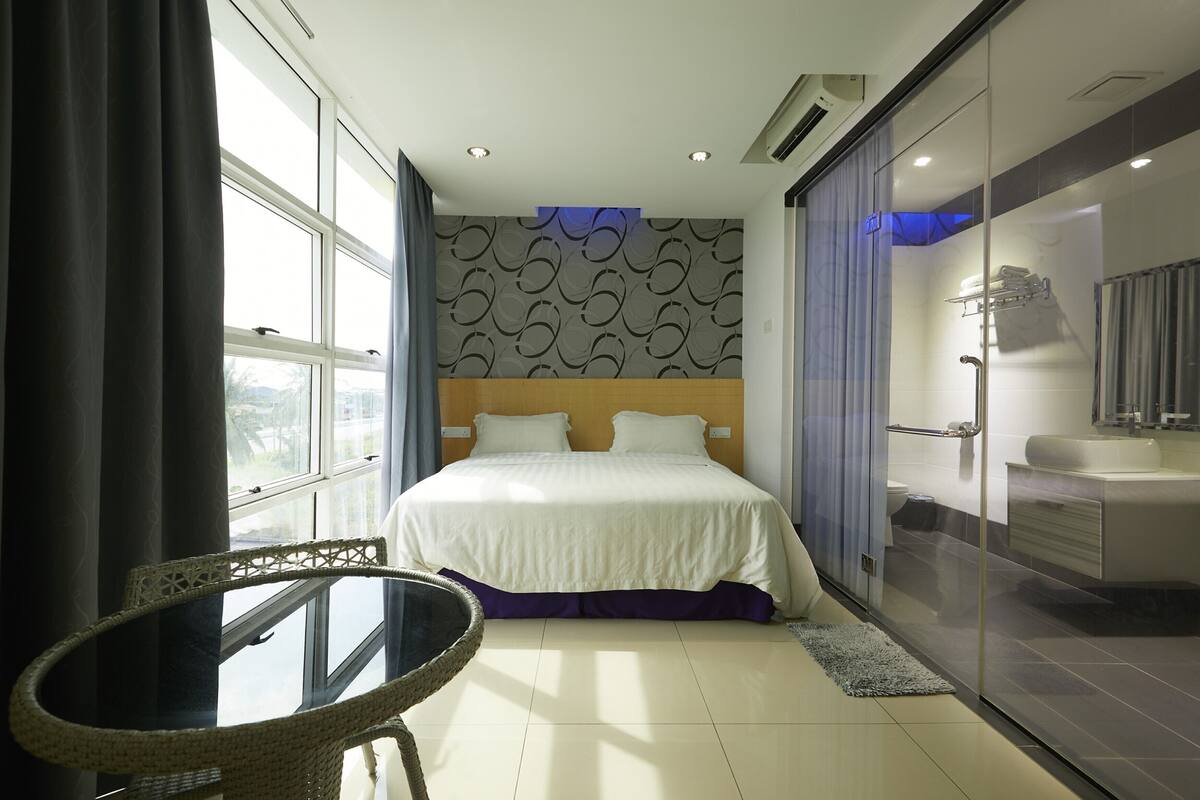 A hotel room with a large bed, a glass shower enclosure, a dining table, and a view of the outdoors.