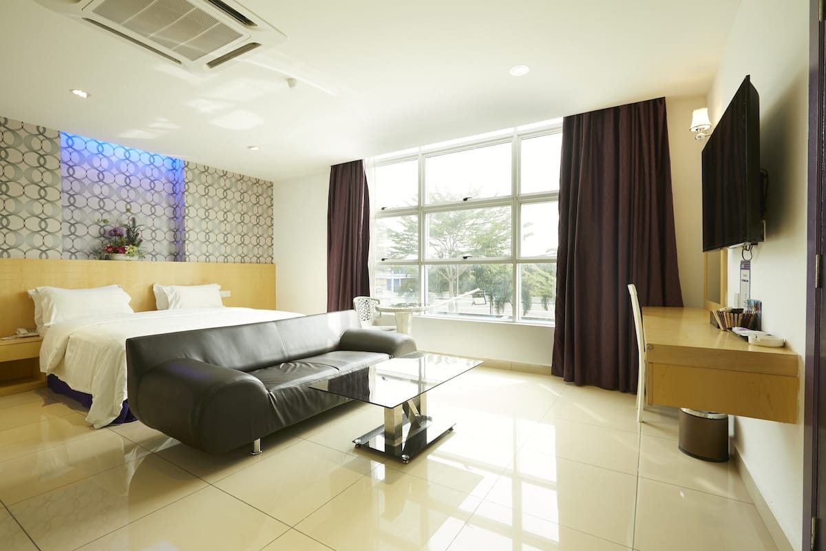 A modern hotel room with a bed, a sofa, a glass coffee table, a desk, and a large window with curtains.