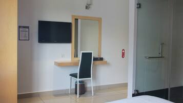 Een hotelkamer met een bed, een bureau met stoel, een aan de muur bevestigde televisie, een spiegel en een douchecabine met glazen deur.