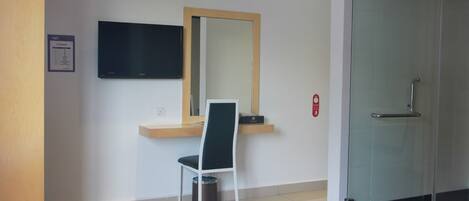 A hotel room with a bed, a desk with a chair, a TV mounted on the wall, a mirror, and a glass shower door.