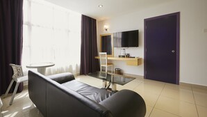 Ett modernt hotellrum med ett matbord med glasskiva, en soffa, en stol, en platt-TV som är monterad på väggen och en dörr.