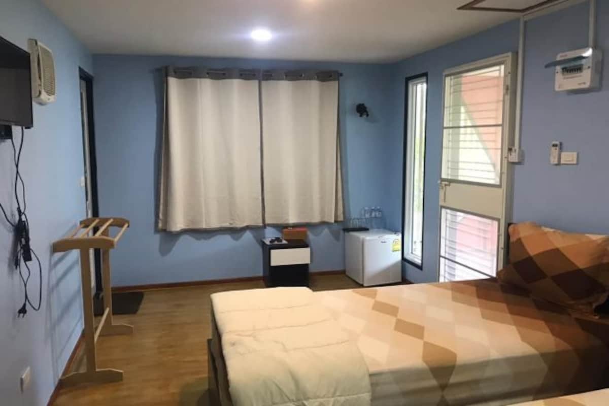 A bedroom with a bed, a desk, a chair, a window with curtains, a bedside table, and a wall-mounted TV.