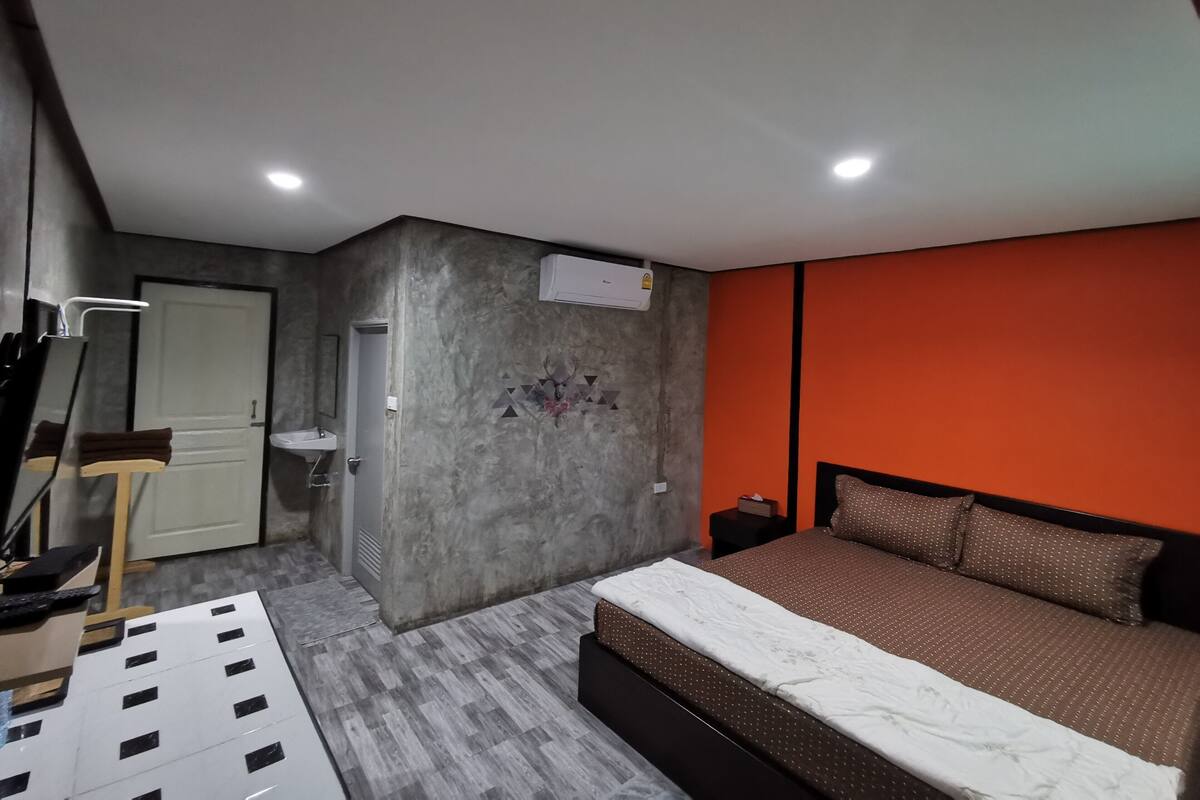 A bedroom with a bed, a desk, and a chair. There is a door and a wall-mounted air conditioner.