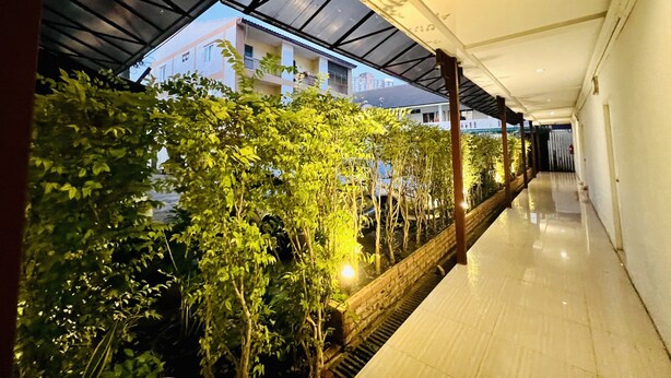 A corridor with a glass ceiling, a brick planter with greenery, and a view of a building in the background. - INN Kham Apartment (Chiang Mai)
