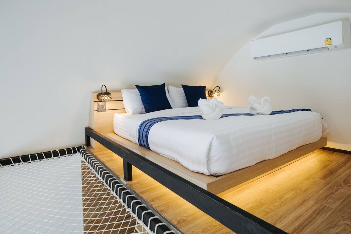 Ein ordentlich bezogenes Bett mit weißer und blauer Bettwäsche, ein Nachttisch mit Lampe sowie eine Wand-Klimaanlage.