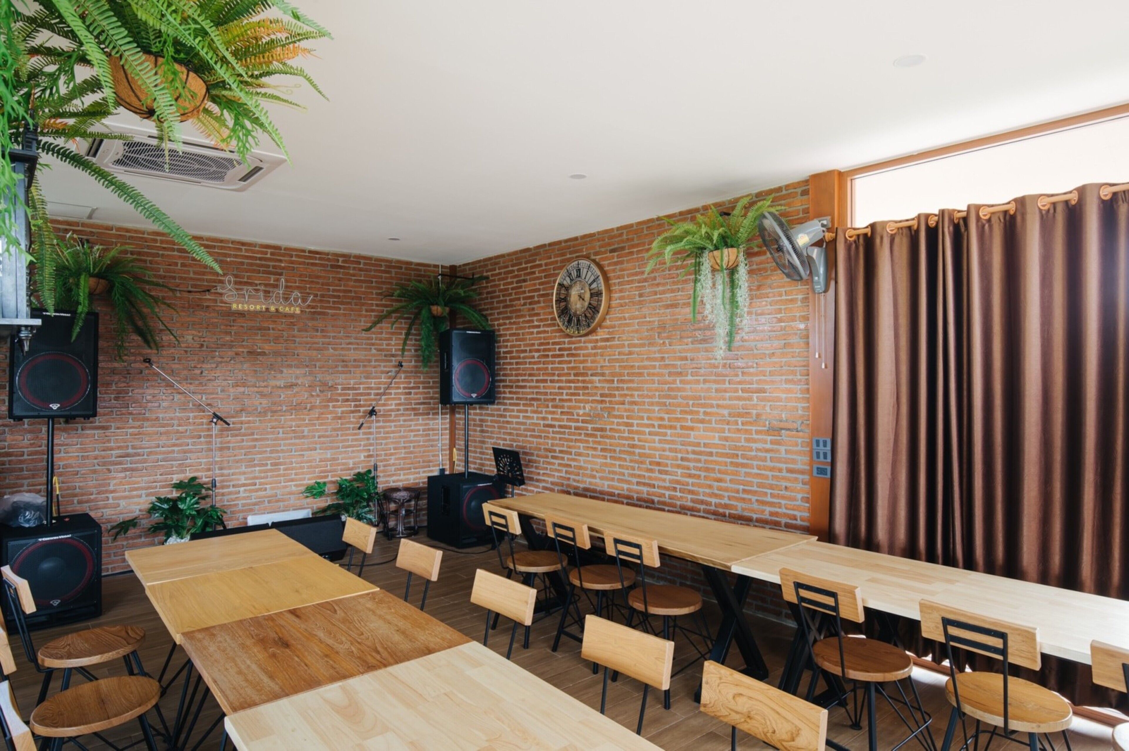 Una habitación amplia con pared de ladrillo, mesas y sillas de madera, plantas en macetas y un reloj.