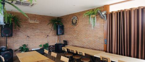Una habitación amplia con pared de ladrillo, mesas y sillas de madera, plantas en macetas y un reloj.