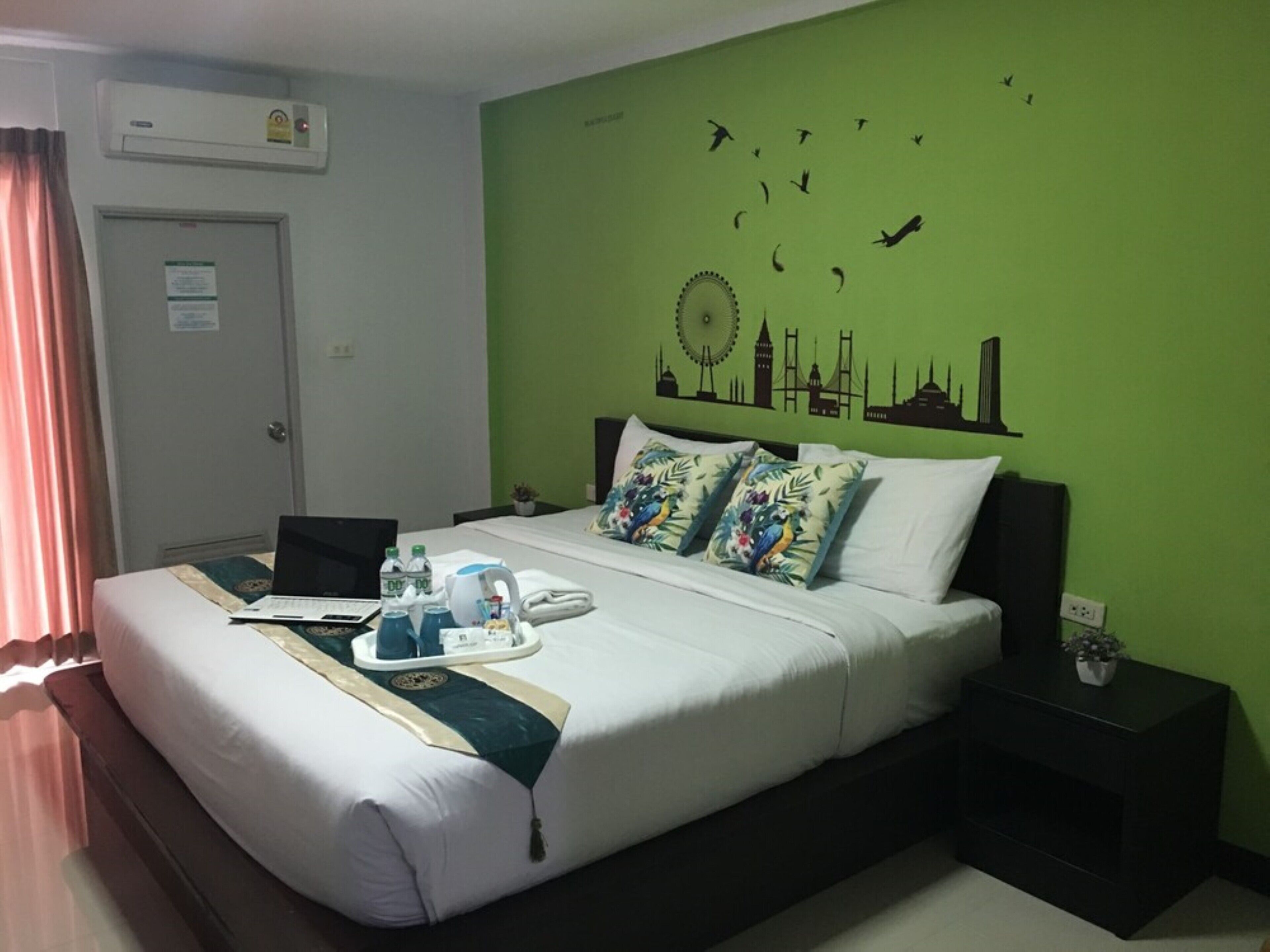 A hotel room with a green wall, a bed with white bedding, a desk with a laptop, and a nightstand with a plant.