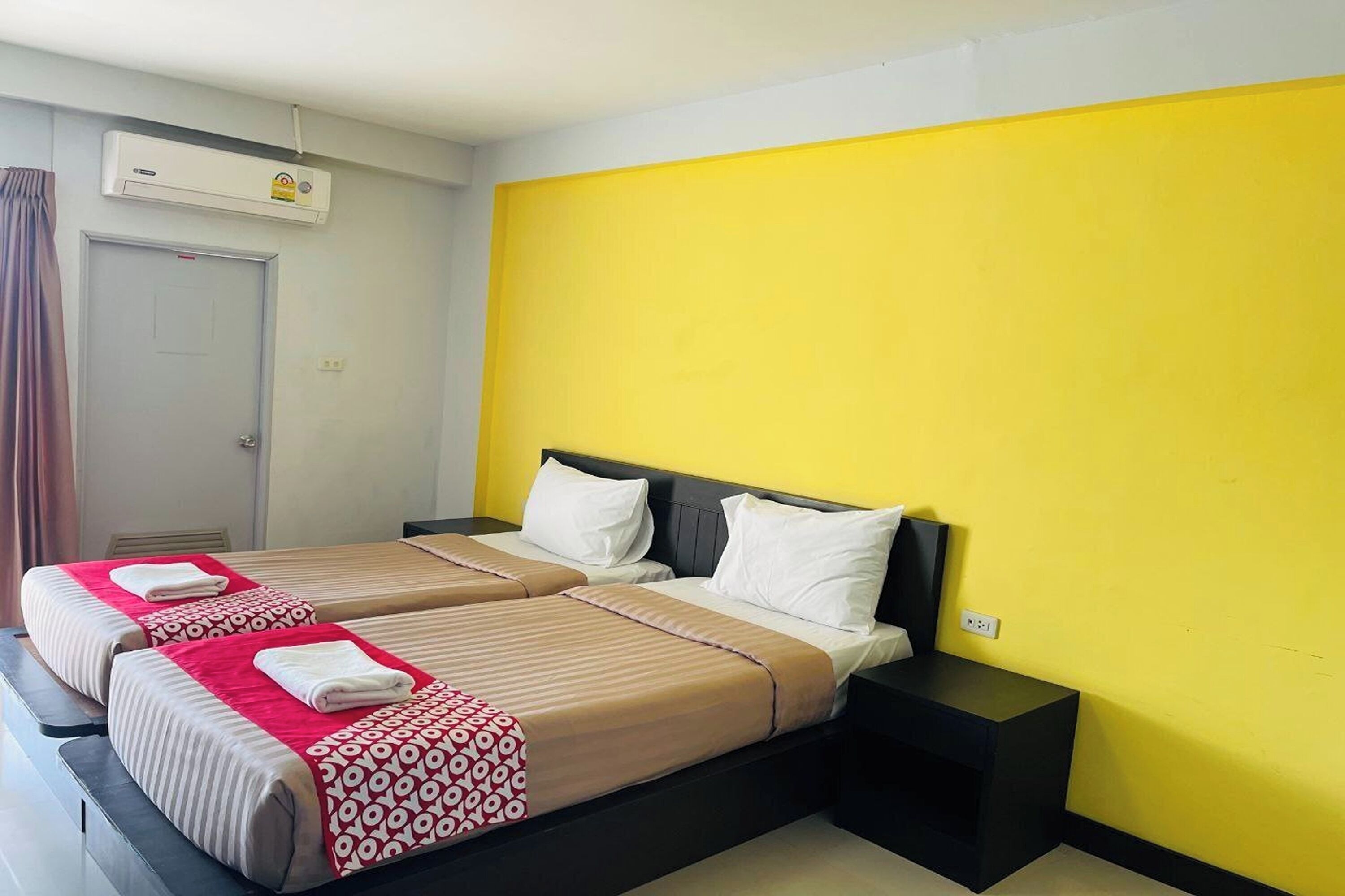 A hotel room with two beds, a yellow accent wall, and a white air conditioning unit.