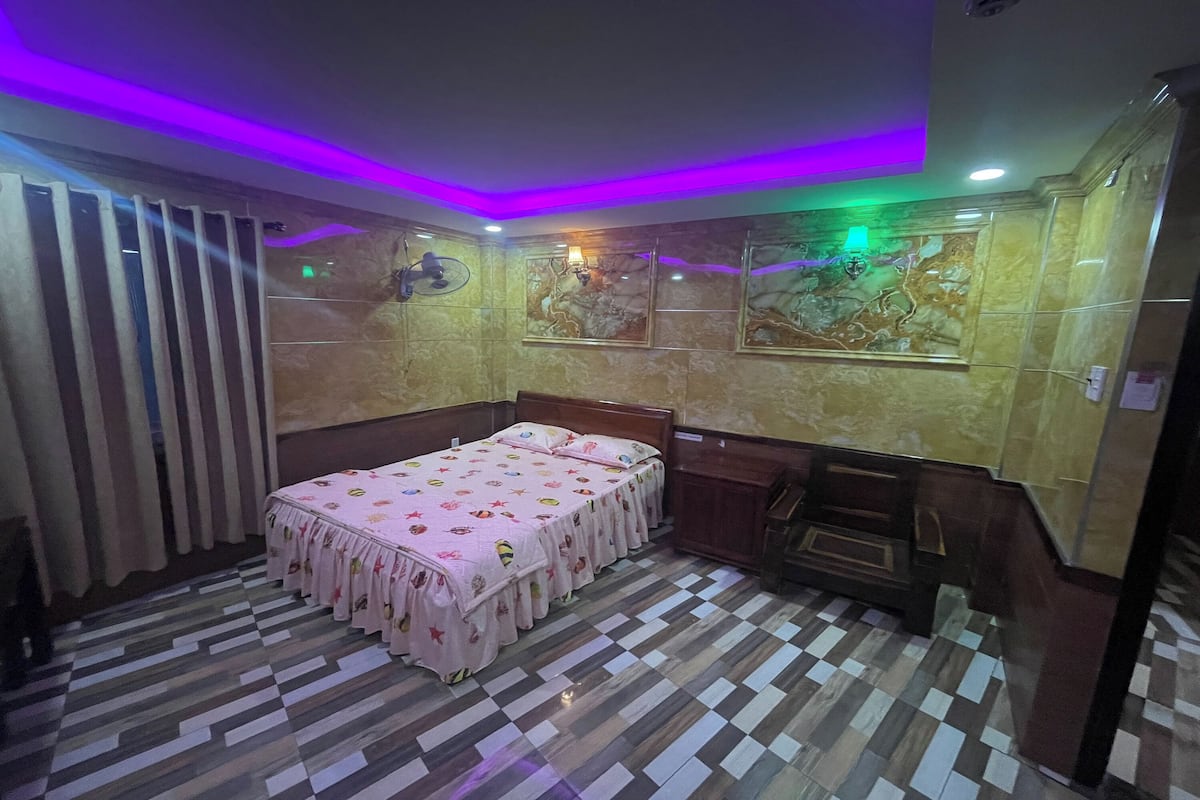 A hotel room with a checkered floor, a bed with a pink bedspread, and a wall-mounted fan.