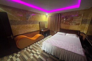 Hotelkamer met een geruit vloerkleed, een bed met witte dekbedovertrek, een oranje stoel, een bureau met spiegel en een aan de muur bevestigde televisie.