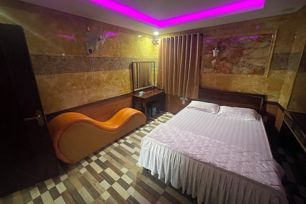 Habitación de hotel con piso a cuadros, cama con cubrecama blanco, sofá naranja, escritorio con espejo e iluminación ambiental morada.