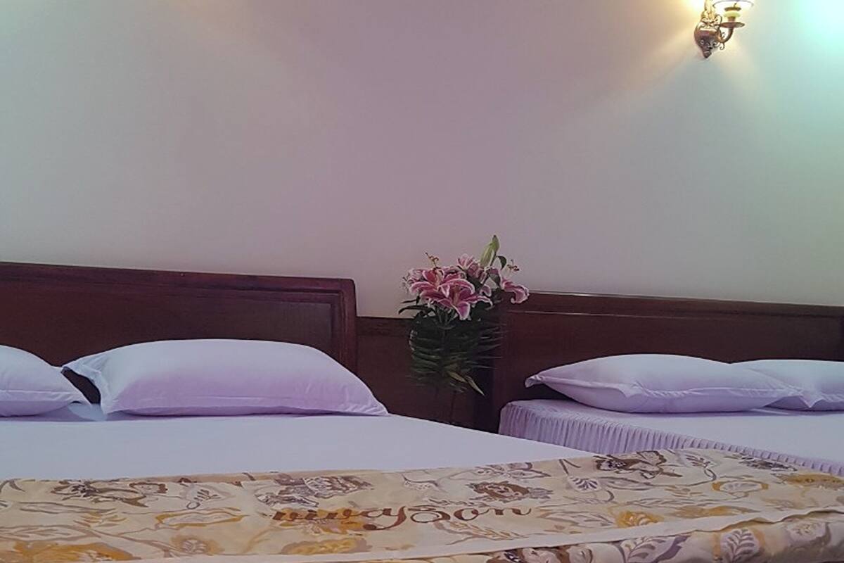 A hotel room with two beds, each with a wooden headboard and white bedding. There are wall-mounted light fixtures and a vase with flowers on a nightstand.