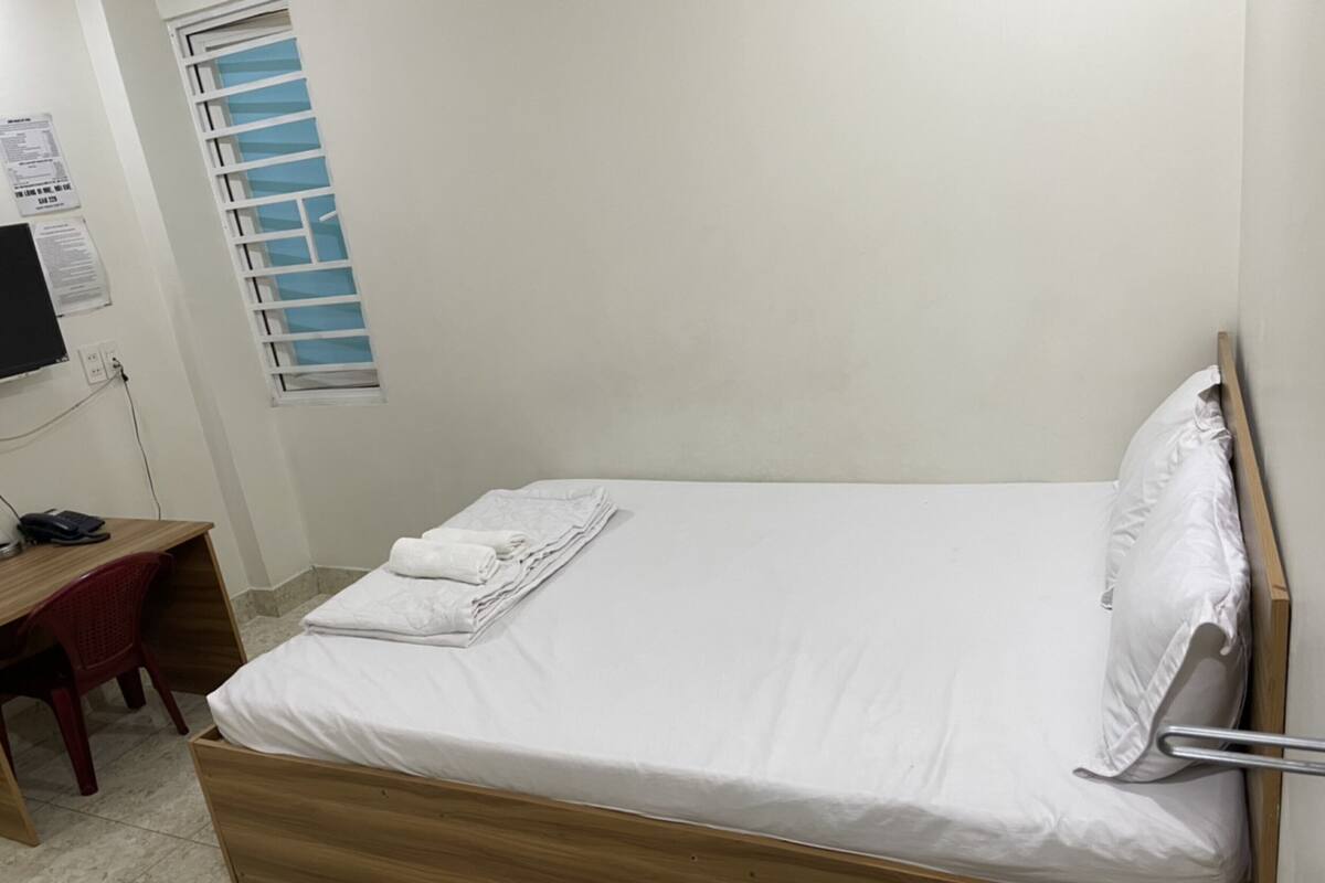 A single bed with a wooden frame, a bedside table with a chair, and a window with blinds.