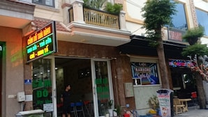 Un edificio con cafetería y bar de karaoke en la planta baja, un hombre de pie afuera y plantas en macetas al frente.