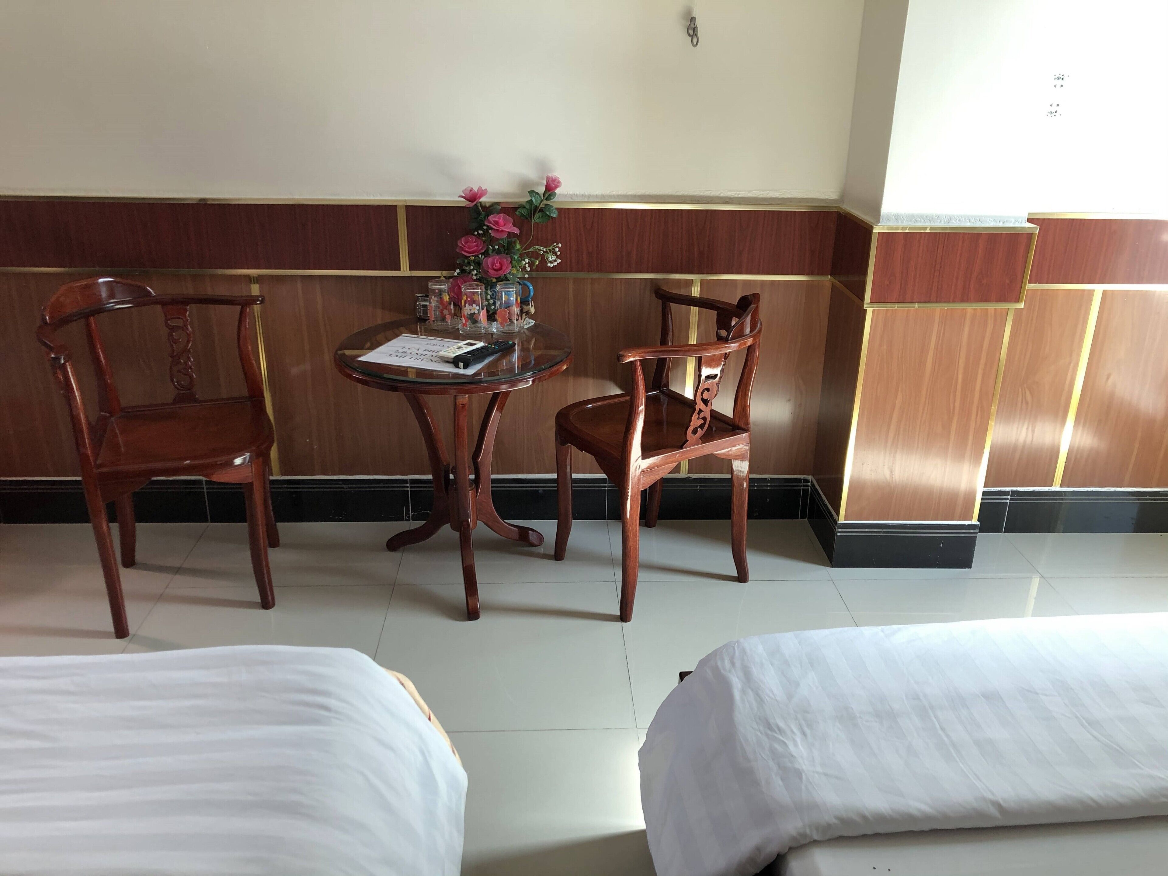 Una habitación de hotel con dos camas individuales, una silla de madera, una mesita redonda con un florero y un control remoto sobre la mesa.