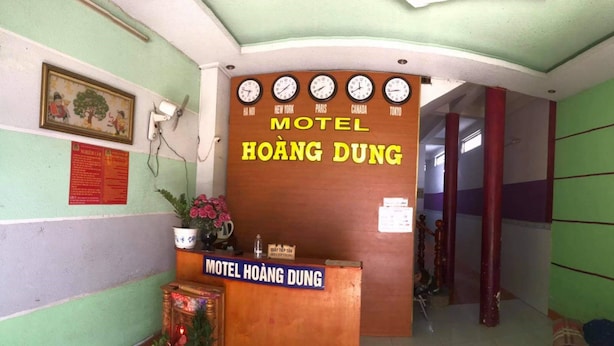 Een receptie met een houten balie, klokken die de tijd in verschillende steden weergeven, en een bord met de tekst 'Motel Hoàng Dung'.