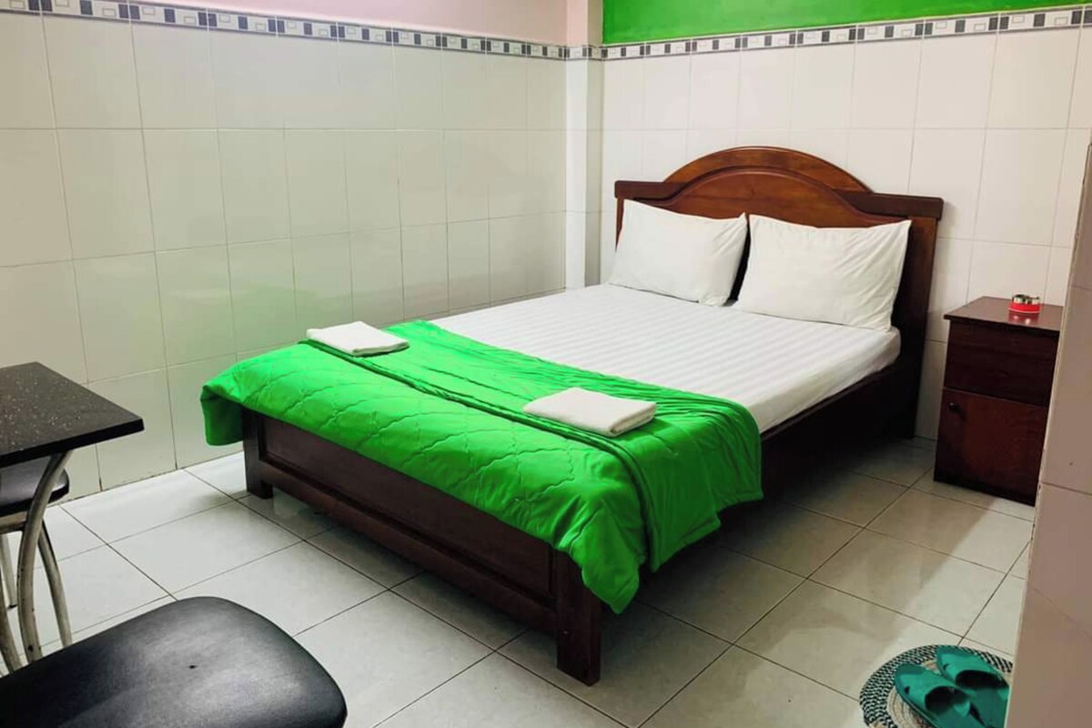 Un lit simple avec une tête de lit en bois, une couvre-lit verte et des oreillers blancs. Il y a une petite table de chevet en bois sur laquelle se trouve un objet rouge. Une chaise est placée devant le lit, et une paire de pantoufles vertes est posée sur le sol.