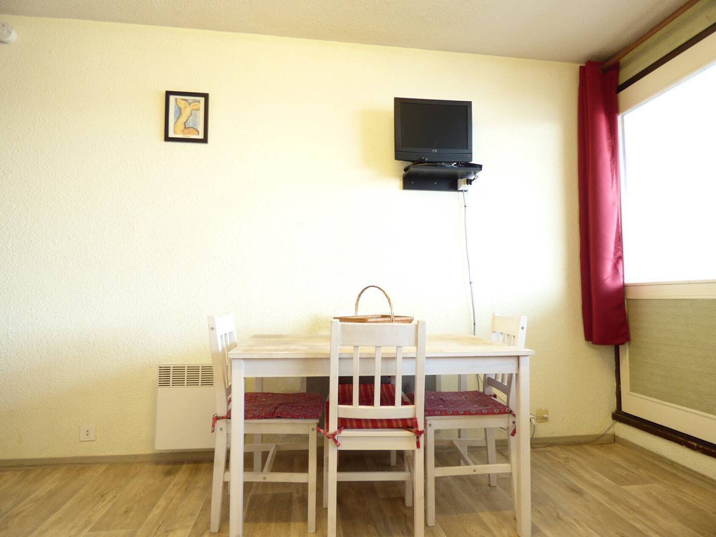 Un pequeño comedor con una mesa y sillas blancas, un televisor montado en la pared y una ventana con cortina roja.