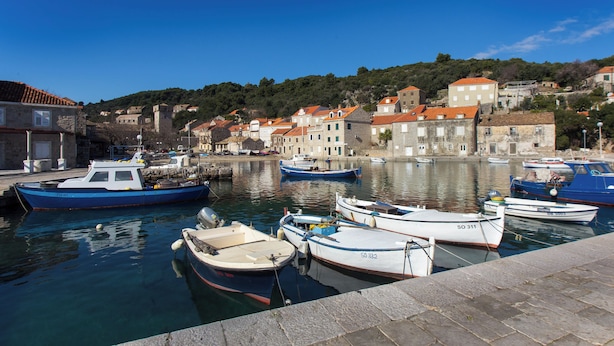 A harbor with boats docked, stone buildings with red roofs, and a hilly backdrop. - Apartment Mali Skoj (Dubrovnik)