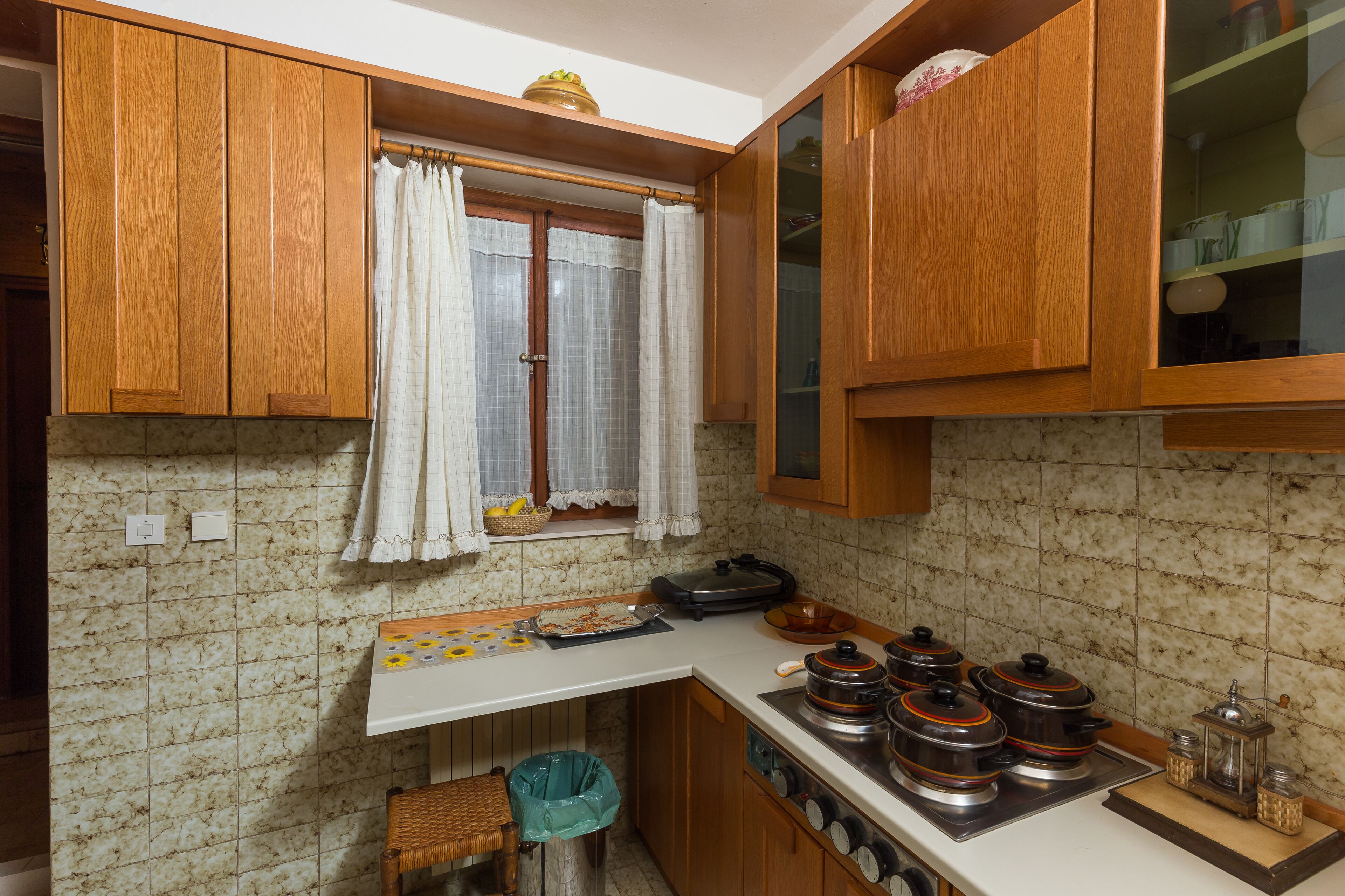 Una cocina con armarios de madera, una ventana con cortinas, salpicadero de azulejos y una estufa con ollas.