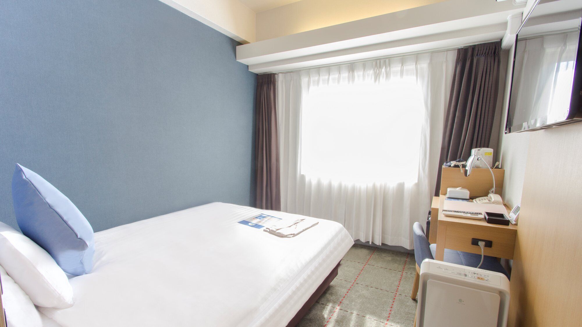 A hotel room with a bed, a desk with a computer, a chair, a window with curtains, and a bedside table.