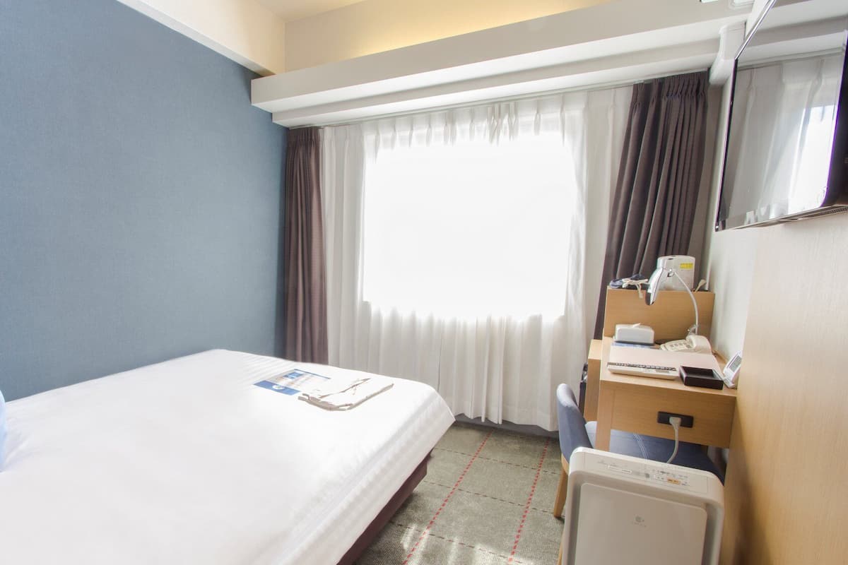 Ein Hotelzimmer mit einem Bett, einem Schreibtisch mit Computer, einem Stuhl, einem Fenster mit Vorhängen und einem Nachttisch.