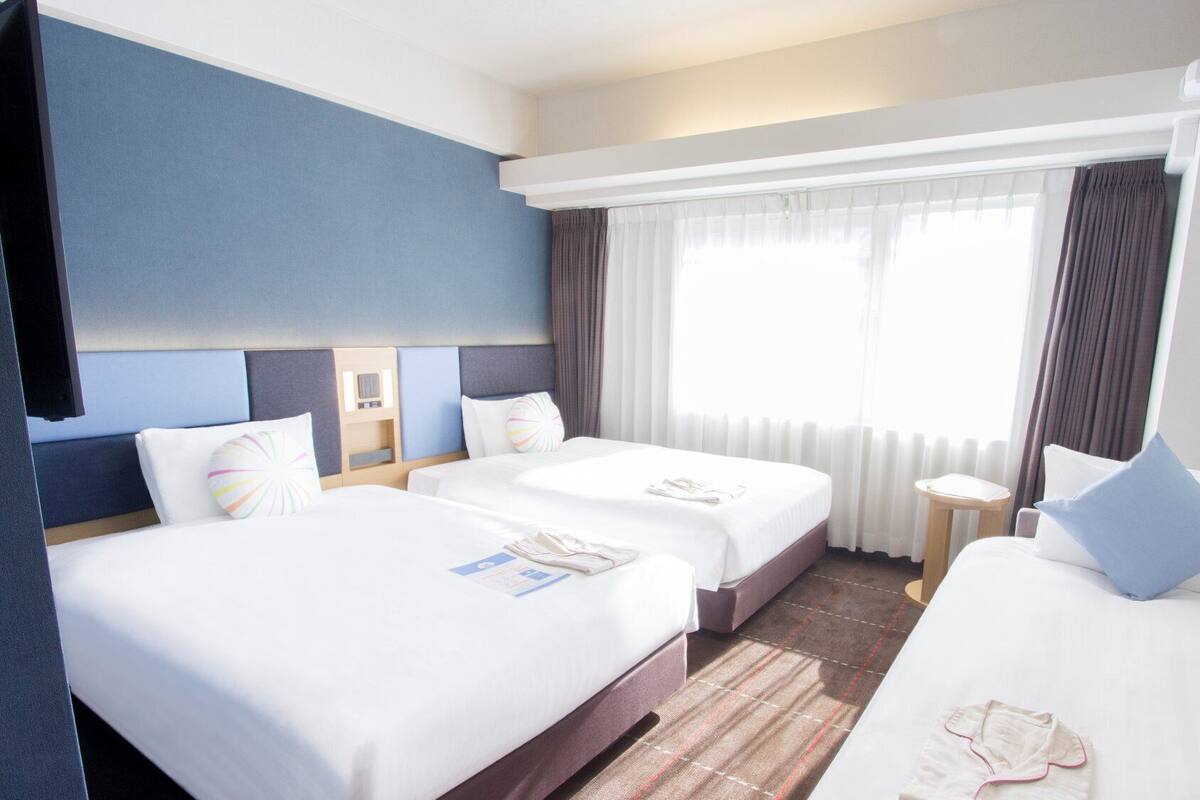 Ein Hotelzimmer mit zwei Betten, einem Fernseher, einem kleinen Tisch und einem Fenster mit Vorhängen.