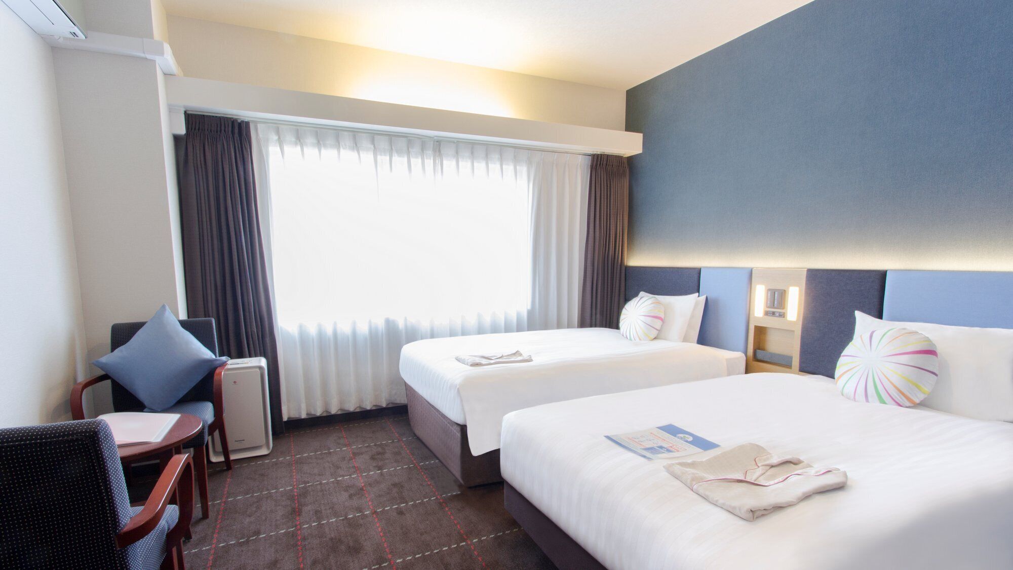 Ein Hotelzimmer mit zwei Betten, einem Schreibtisch, einem Stuhl und einem Vorhangfenster.