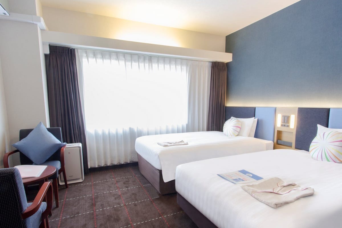 Ein Hotelzimmer mit zwei Betten, einem Schreibtisch, einem Stuhl und einem Vorhangfenster.
